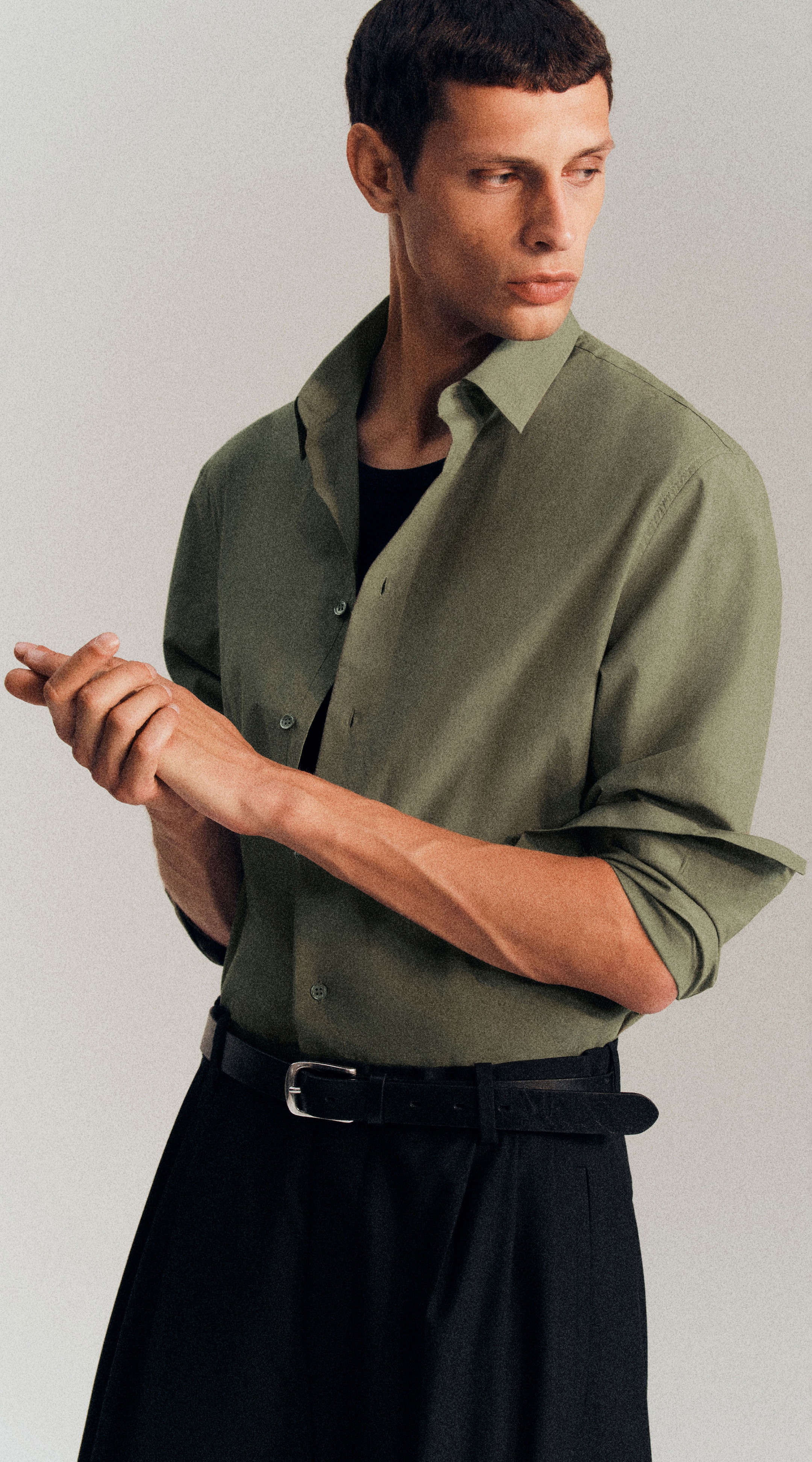 Man in a green button-up shirt and black pants, looking to the side, adjusting his wrist, belt visible, against a plain background.