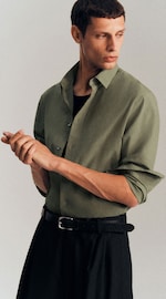 Placeholder for: Man in a green button-up shirt and black pants, looking to the side, adjusting his wrist, belt visible, against a plain background.