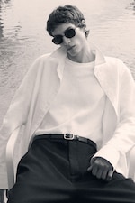 Placeholder for: Person wearing sunglasses and a white open-collared shirt, reclining with a casual pose in front of a reflective water background.