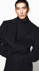 Placeholder for: Close-up portrait of a man wearing a dark wool overcoat and ribbed turtleneck, hands in pockets against a white background.