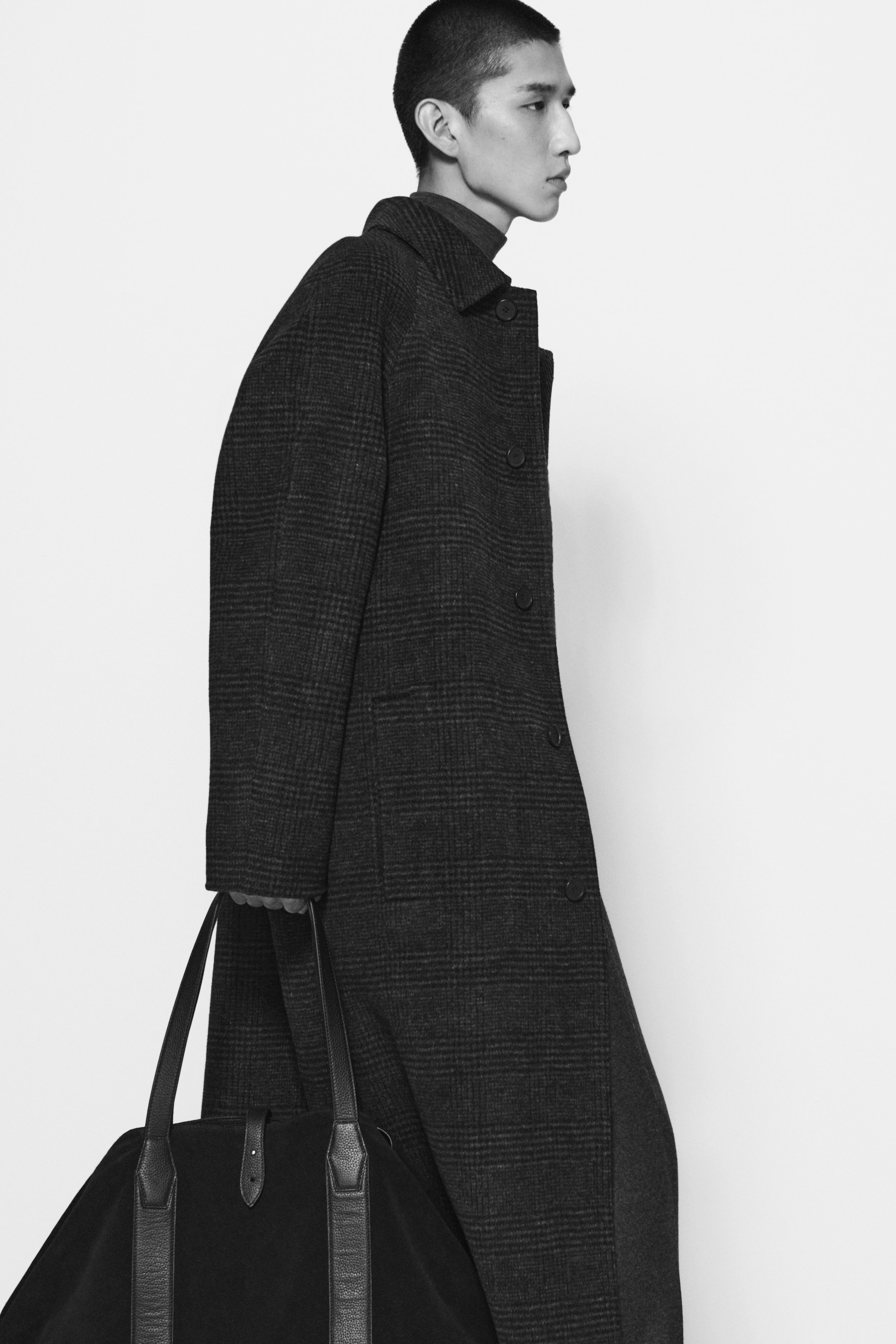 Person in a long, dark plaid coat carrying a large black bag, walking against a plain white background.