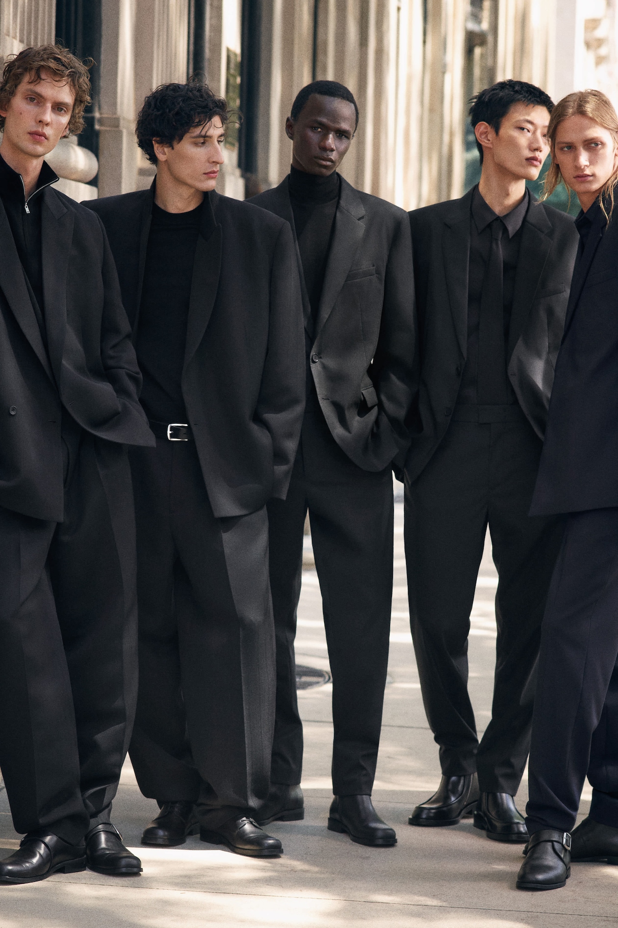 Five people in black suits stand together on a city sidewalk, gazing in different directions. The scene is stylish and urban.