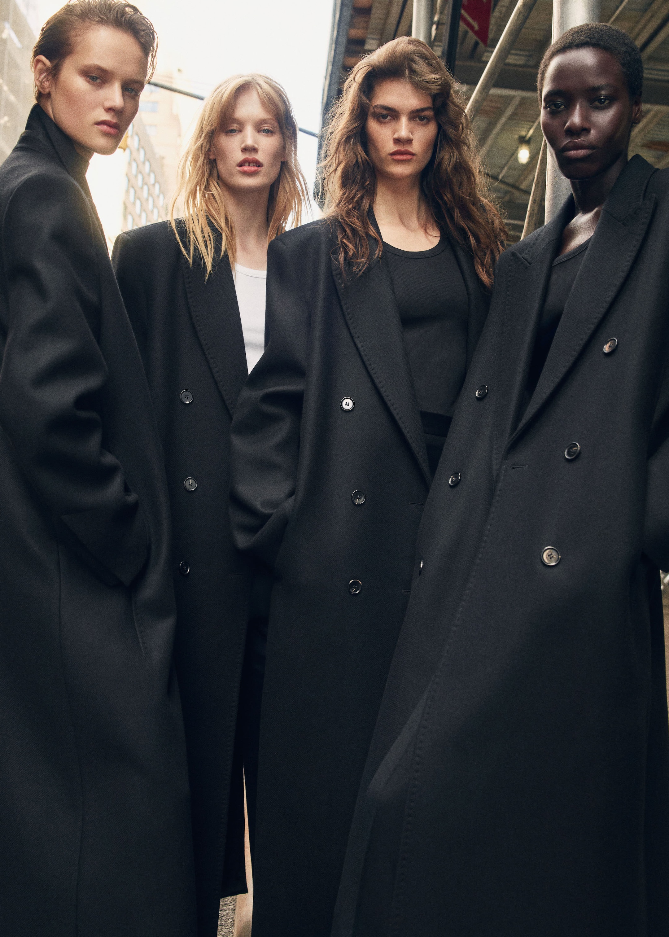 Four models stand confidently on a city street, wearing long, black coats. They pose under scaffolding, exuding a strong and stylish presence.
