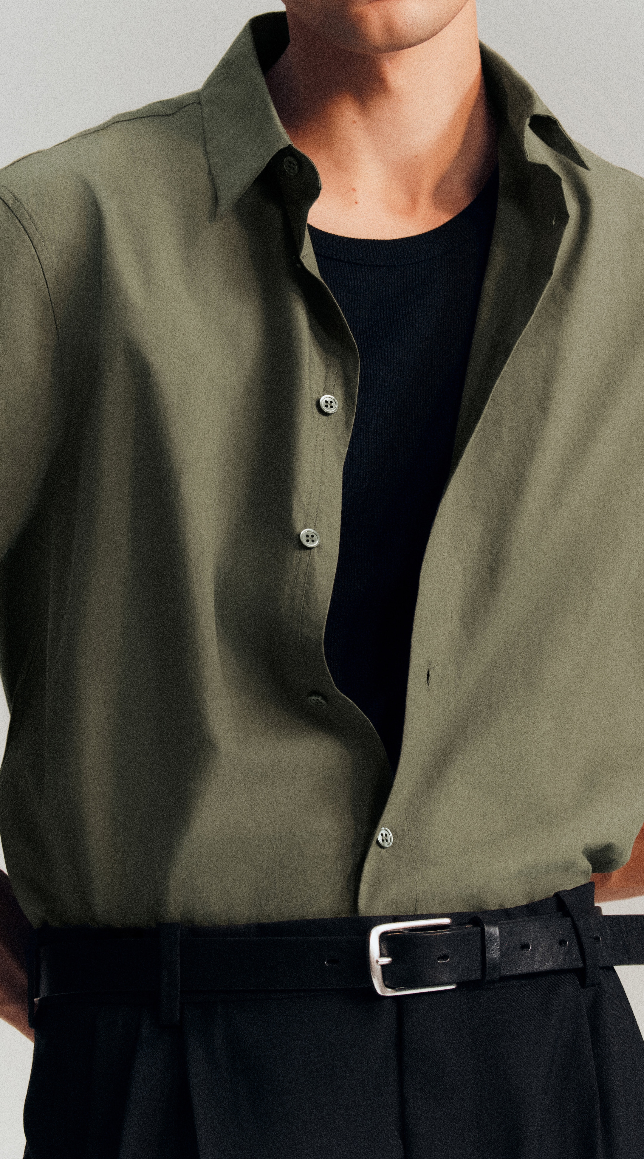 A man wearing a green button-up shirt over a black T-shirt, paired with black trousers and a black belt, standing against a neutral background.