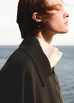 Placeholder for: Profile of a person with windswept hair, wearing a dark coat over a white shirt, set against a blurred ocean background.