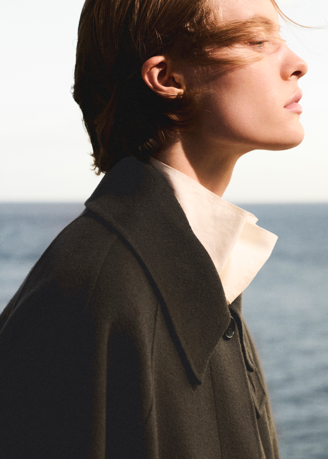 A person wearing a dark coat and white shirt stands in profile with the ocean in the background, hair gently blowing in the wind.