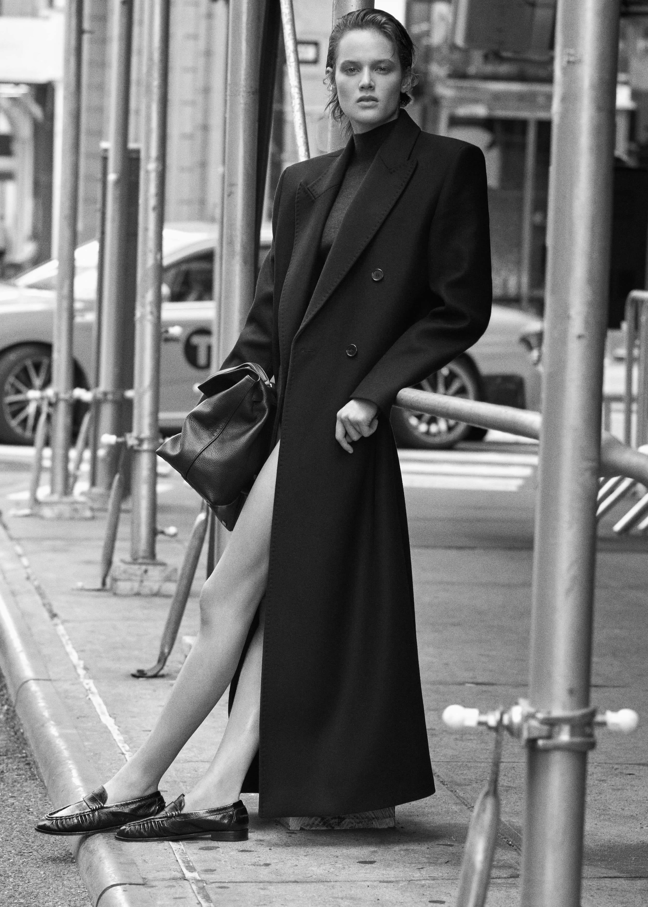 A woman in a long black coat and loafers leans against scaffolding in an urban setting, holding a bag.