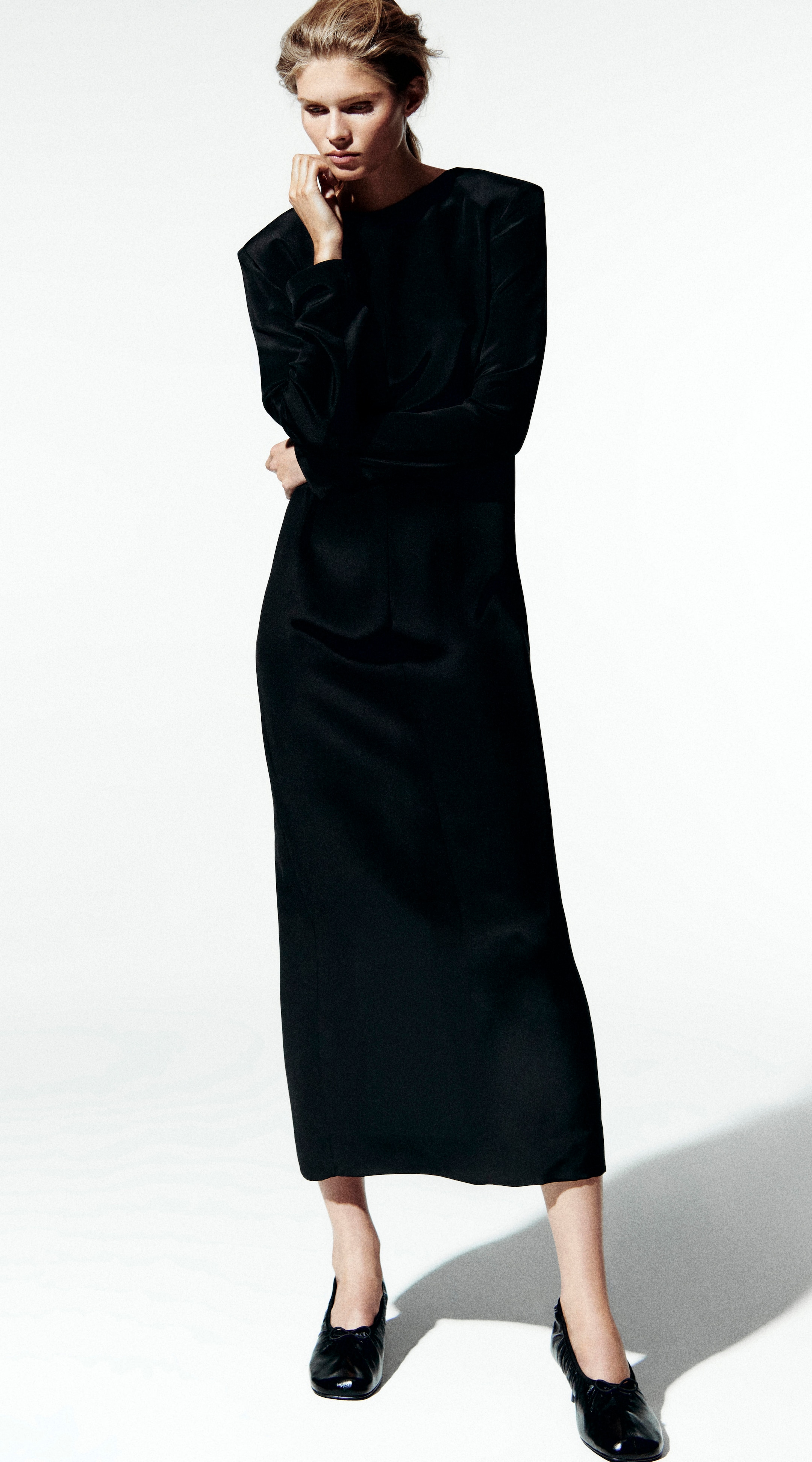 Woman in a long black dress and black shoes with a thoughtful expression, standing against a plain white background.