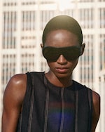 Placeholder for: Person with short hair wearing oversized sunglasses and a dark sleeveless top, standing in front of a blurred building with sunlight behind them.