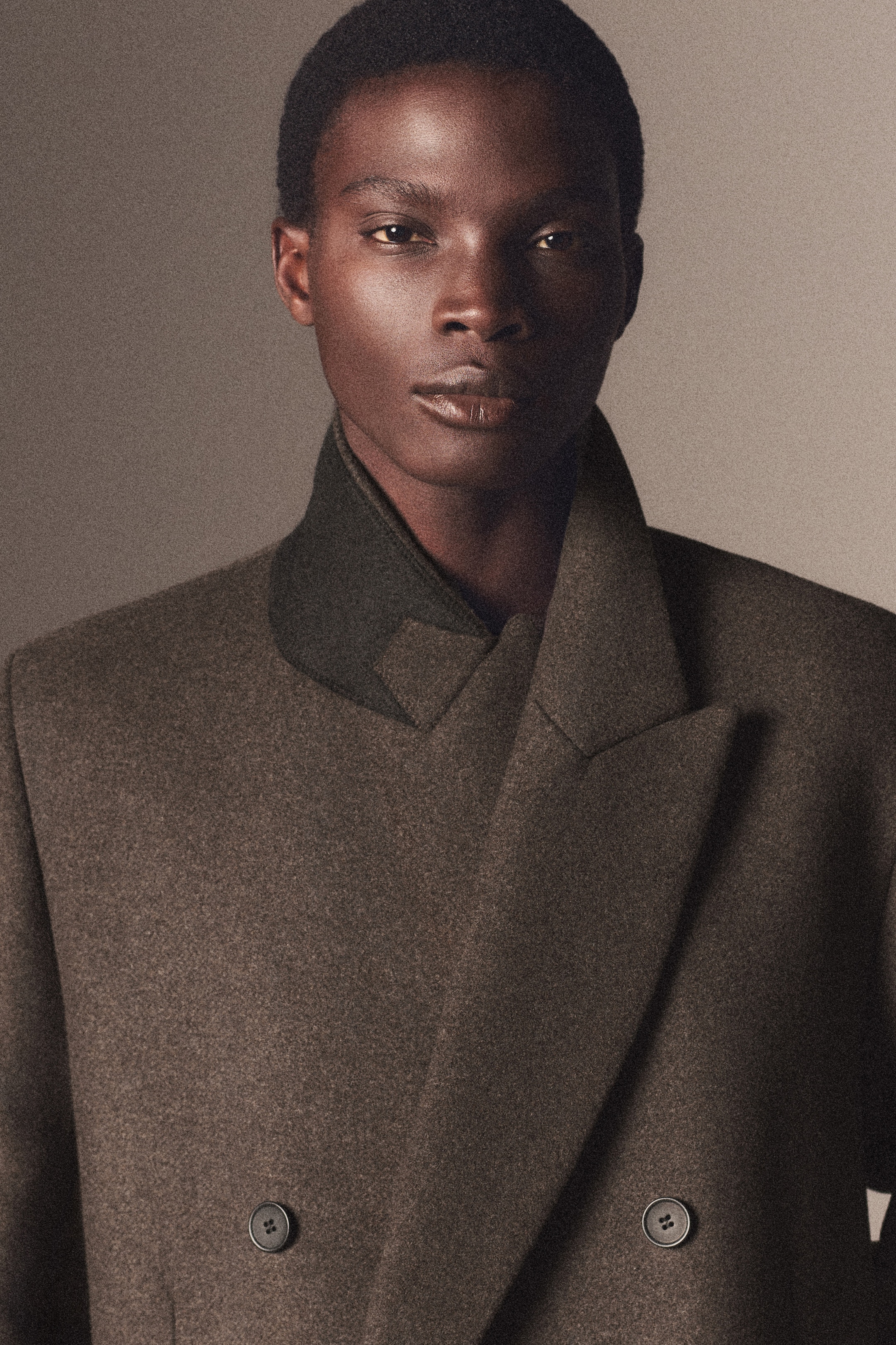 Person in a dark brown double-breasted coat with a contrasting collar, looking directly at the camera against a neutral background.