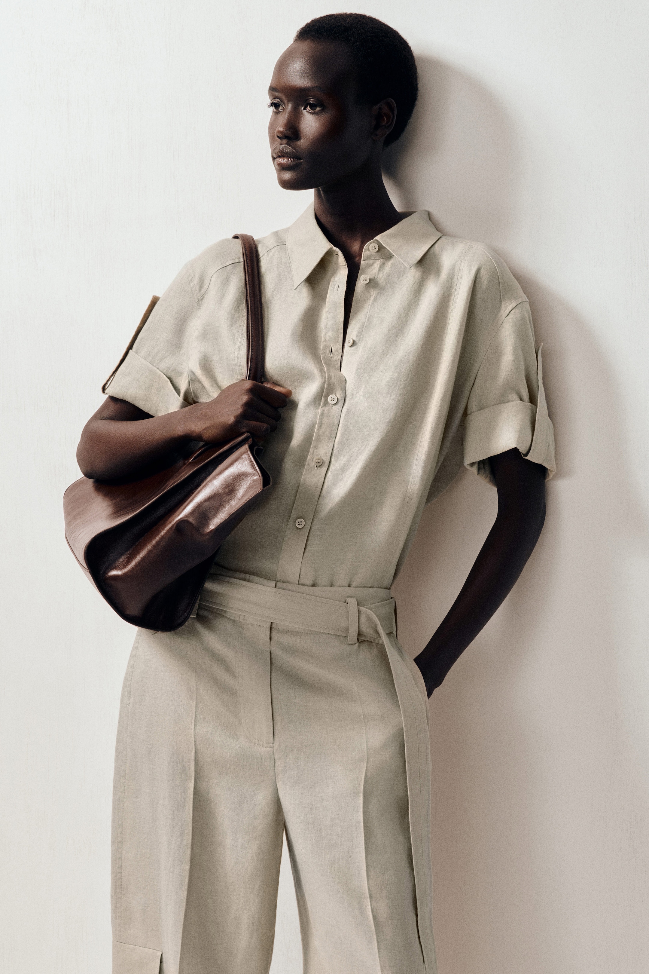 A person in a beige button-up shirt and trousers holds a brown leather bag, standing against a white background.