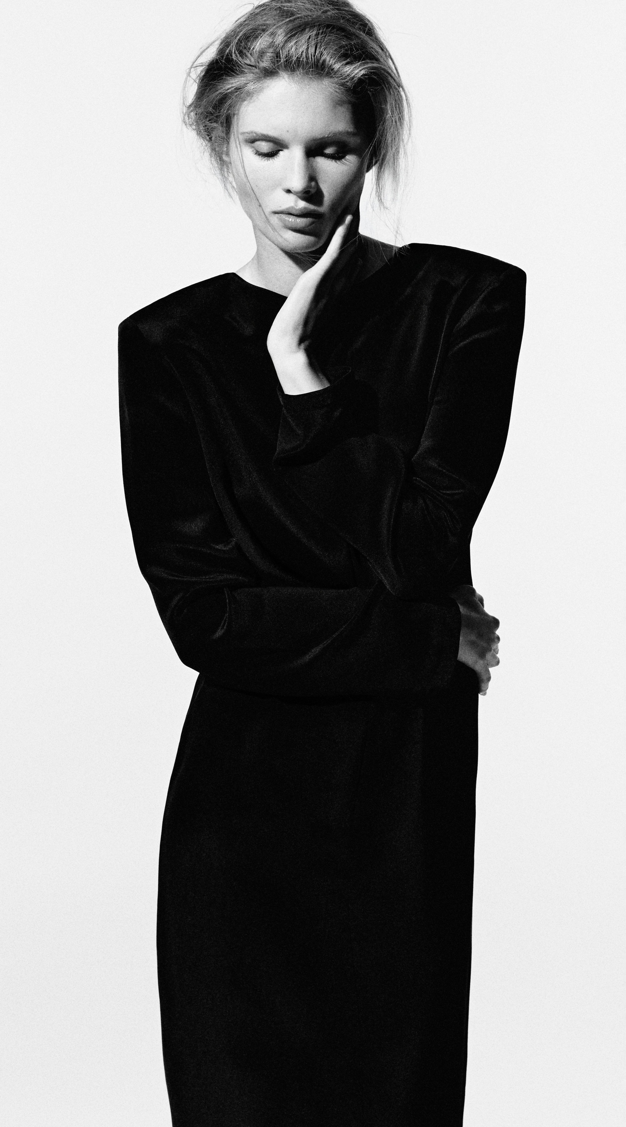 Black and white photo of a woman in a black dress with eyes closed, hand touching her face, exuding elegance and poise.