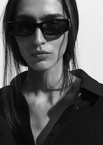 Placeholder for: Person with long hair wearing rectangular sunglasses and a dark shirt, gazing at the camera with a serious expression in black and white.
