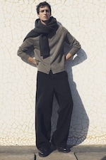Placeholder for: Man posing casually, wearing a gray cardigan, dark scarf, and black wide-leg pants against a textured, light-colored wall.
