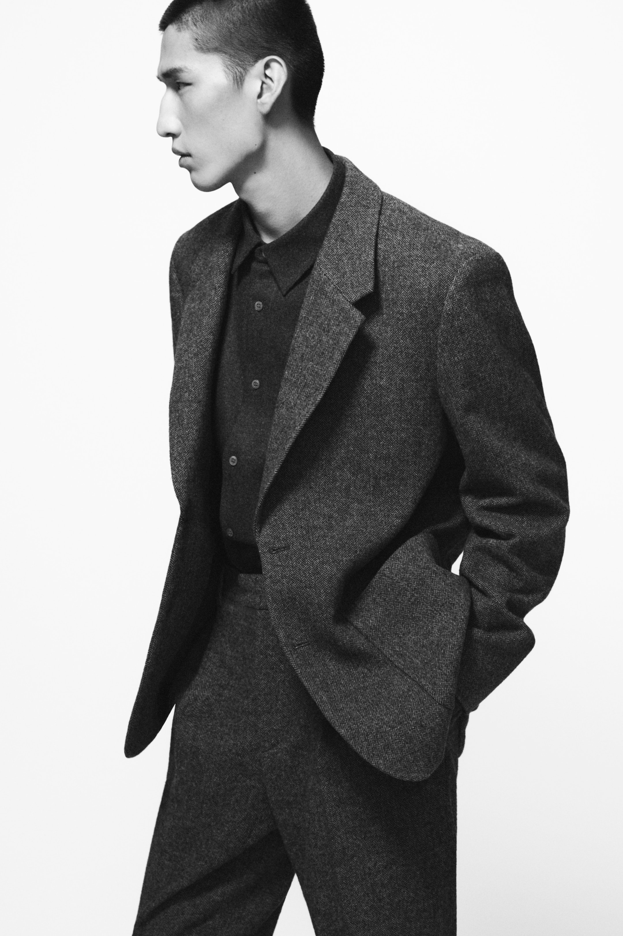 Person in a dark, textured suit with hands in pockets, looking to the side against a plain white background.