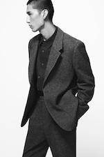 Placeholder for: Person in a dark, textured suit with hands in pockets, looking to the side against a plain white background.