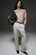 Placeholder for: Model wearing a beige sweater, light-gray tailored trousers, black belt and pointed black shoes, holding a dark brown suede bag.