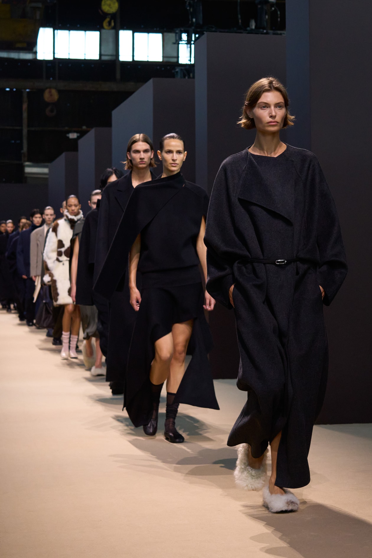 Models walking down a runway, wearing dark, flowing garments and stylish footwear in a fashion show setting.