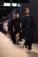 Placeholder for: Models walking down a runway, wearing dark, flowing garments and stylish footwear in a fashion show setting.