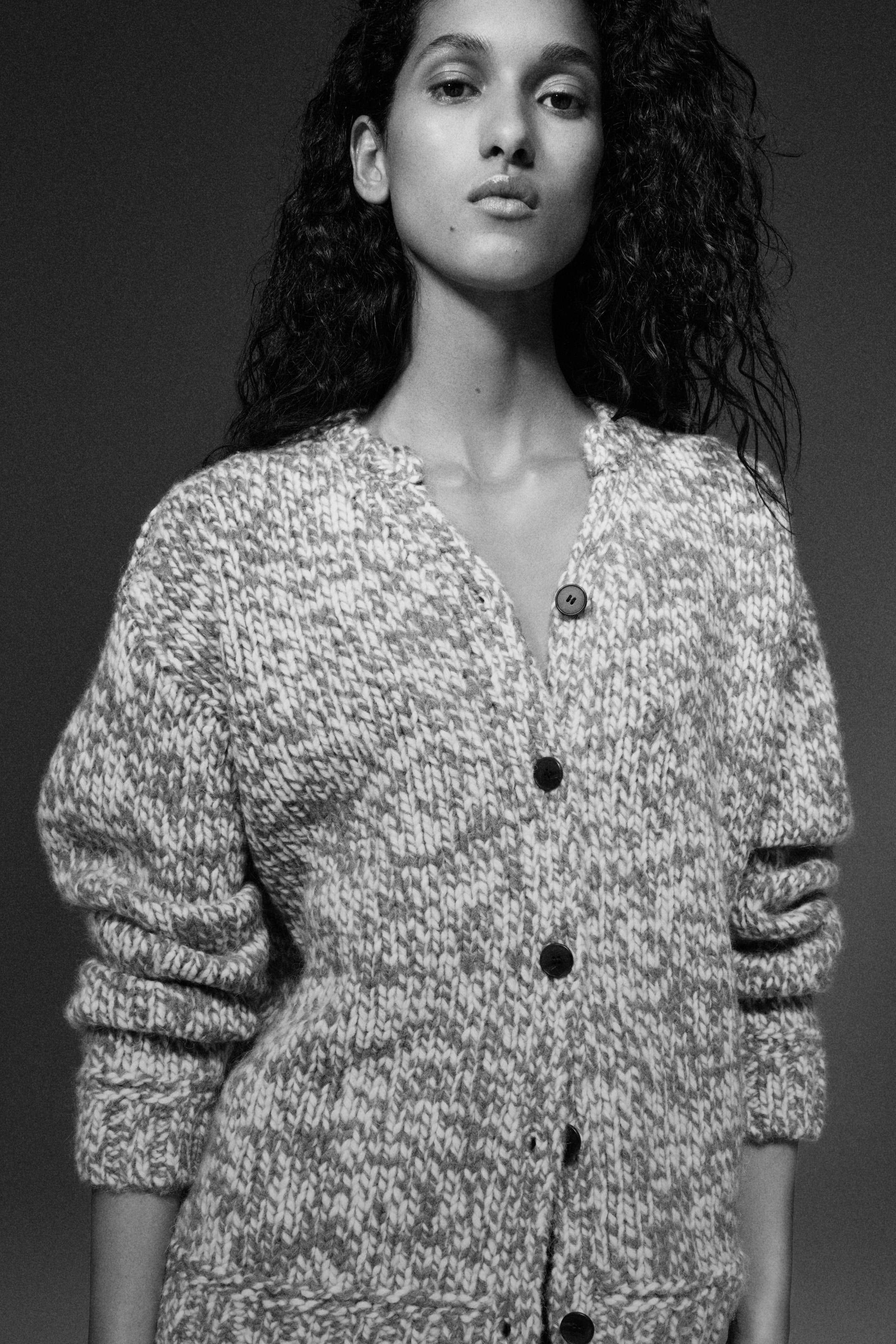 Black-and-white portrait of a woman with long curly hair wearing a chunky marled button-up cardigan, neutral expression.