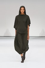 Placeholder for: Model wearing a dark green, loose-fitting outfit with a hood, walking on a white runway.