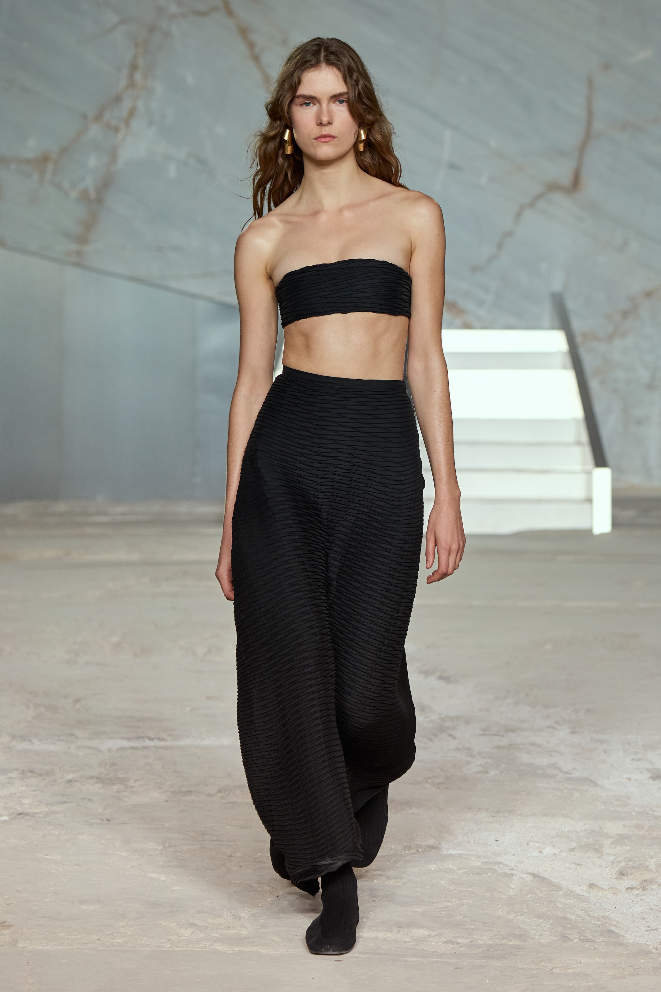 A model walks down a runway wearing a black strapless top and high-waisted wide-leg pants, set against a minimalist marble backdrop.