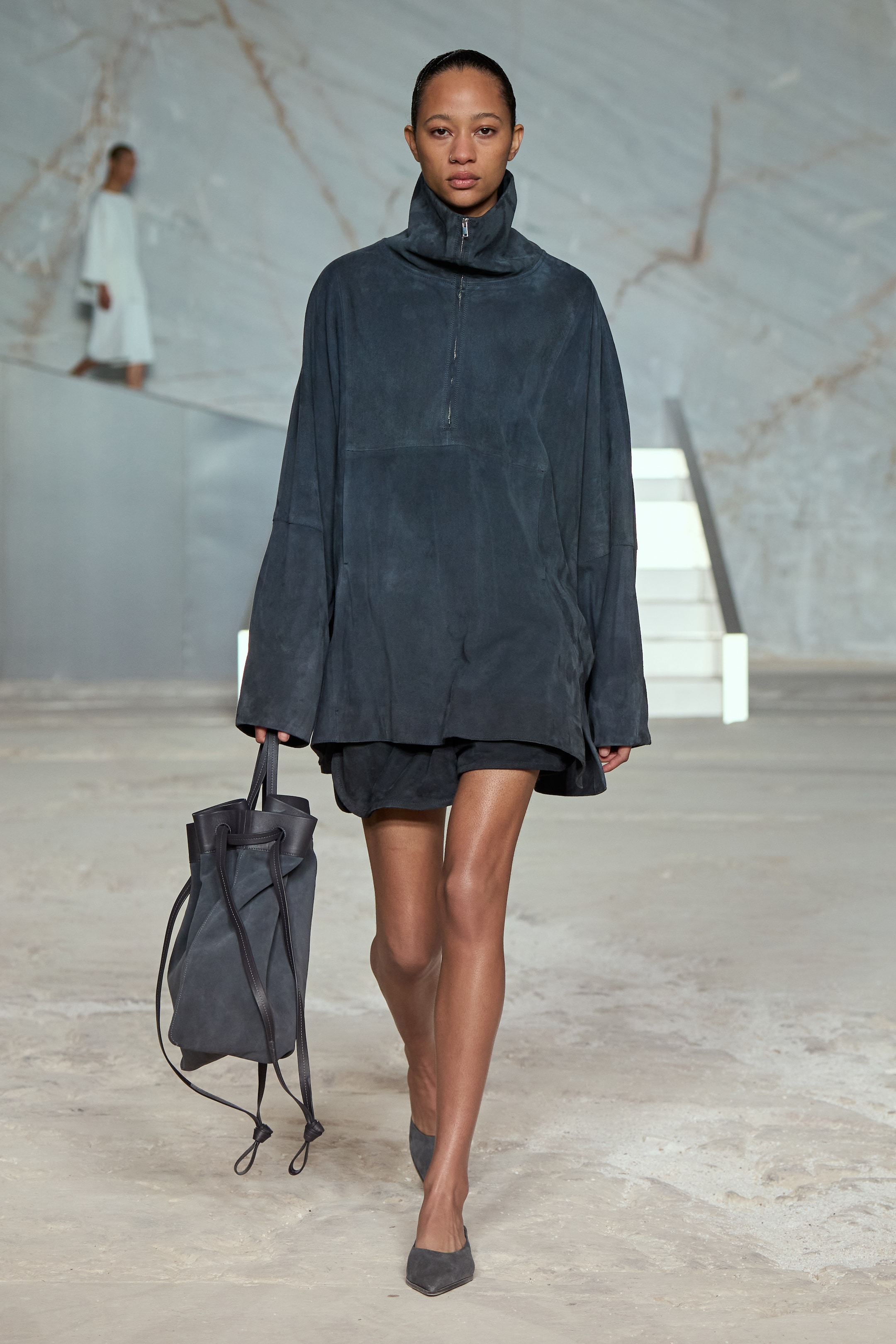 A model in a dark oversized outfit and carrying a matching bag walks on a minimalist runway with a marble backdrop and white stairs.