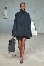 Placeholder for: A model in a dark oversized outfit and carrying a matching bag walks on a minimalist runway with a marble backdrop and white stairs.