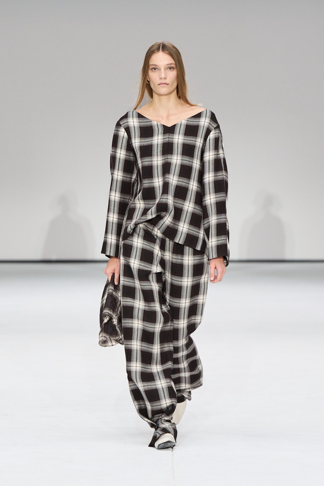 Model wearing a loose-fitting black and white plaid outfit, holding a small bag, walks on a minimalist runway.