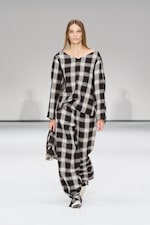 Placeholder for: Model wearing a loose-fitting black and white plaid outfit, holding a small bag, walks on a minimalist runway.