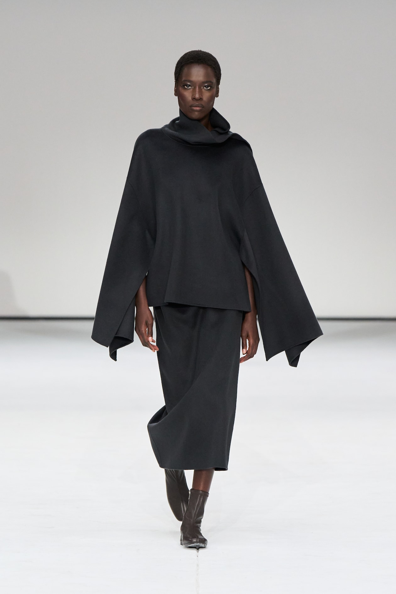 Model in a flowing black outfit with a cowl neck and wide sleeves walks on a white runway, wearing brown ankle boots.