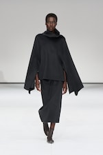 Placeholder for: Model in a flowing black outfit with a cowl neck and wide sleeves walks on a white runway, wearing brown ankle boots.