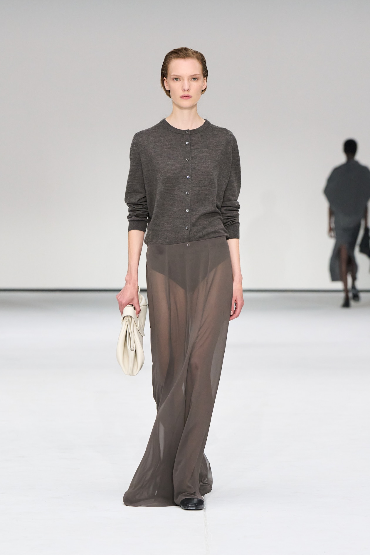 Model in a dark cardigan and sheer skirt walks the runway, holding a white clutch, against a minimalist white background.
