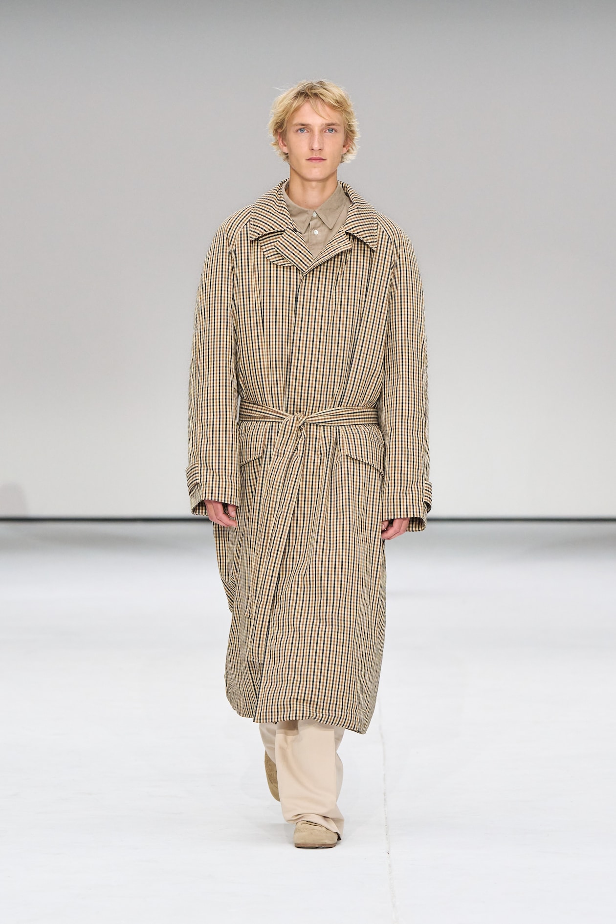 Person walking on a runway wearing a long, plaid coat with a belt, over beige pants and shoes, against a plain background.