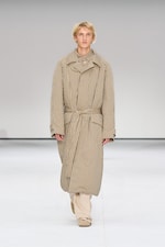 Placeholder for: Person walking on a runway wearing a long, plaid coat with a belt, over beige pants and shoes, against a plain background.
