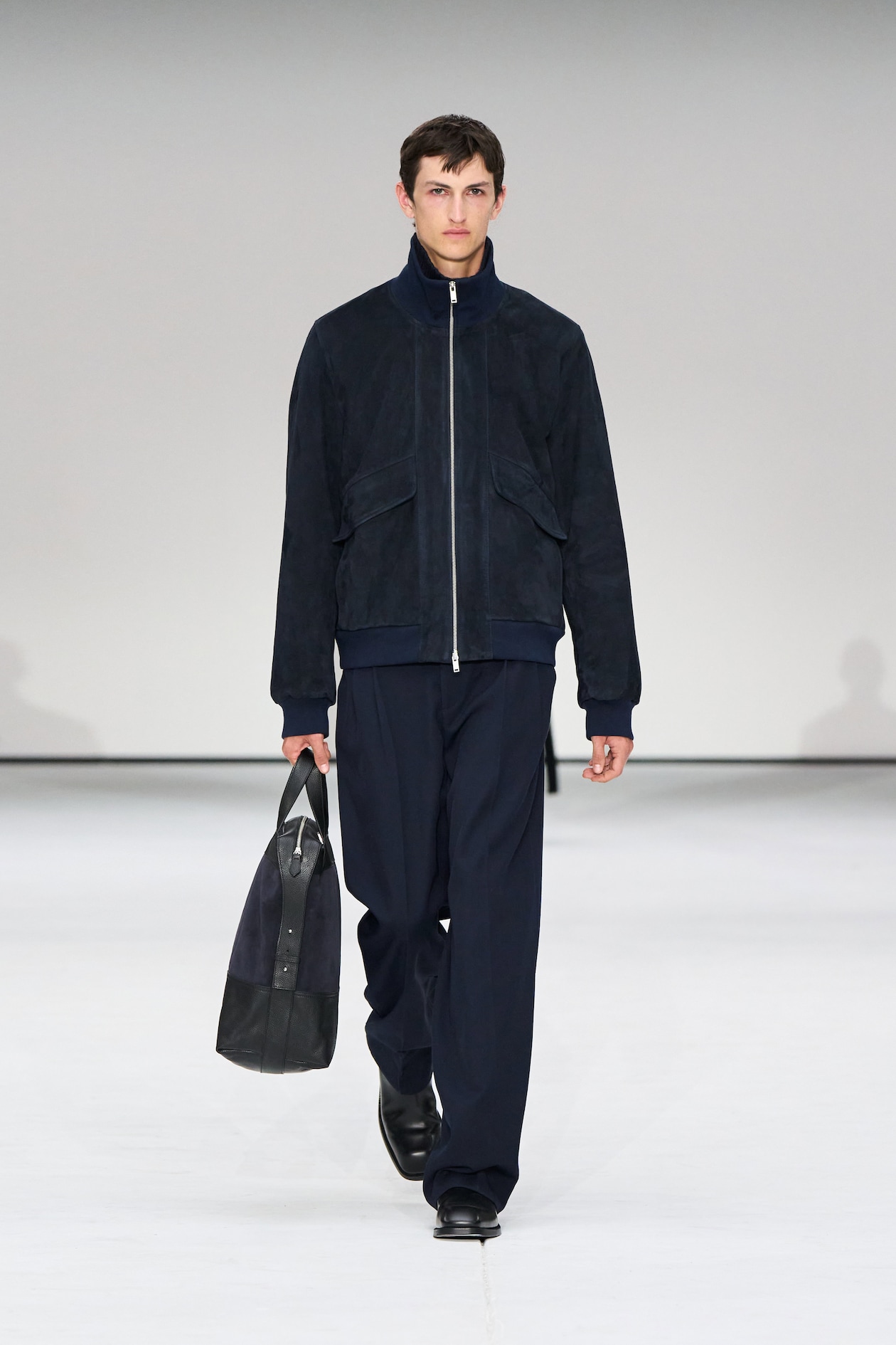 A model in a dark jacket and trousers walks on a runway, holding a large black bag, against a plain background.