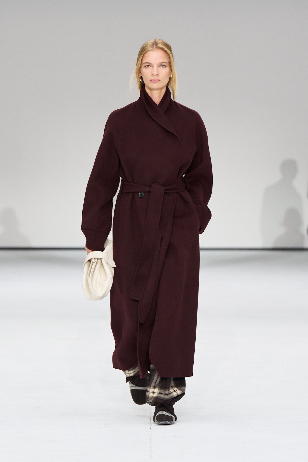 Model in a long, dark maroon coat walks on a runway, holding a white clutch, with a minimalist gray background.
