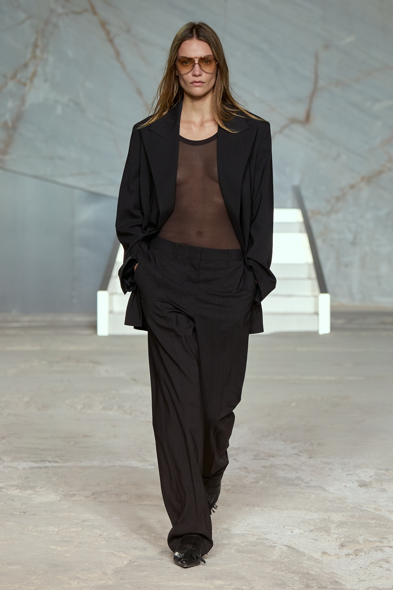 A model in a sheer top, oversized black blazer, and trousers walks confidently on a minimalist runway.