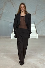 Placeholder for: A model in a sheer top, oversized black blazer, and trousers walks confidently on a minimalist runway.