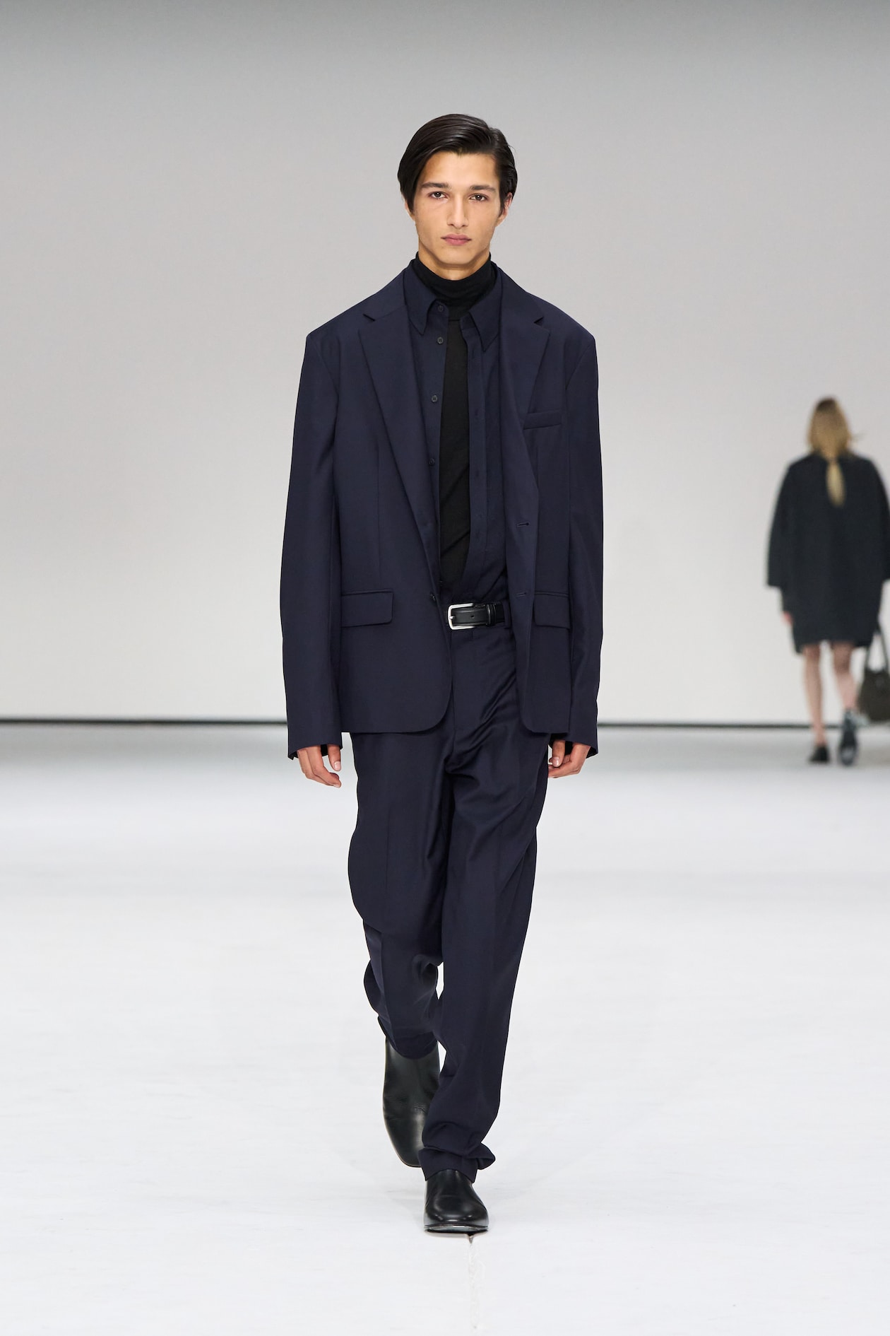 A model in a dark suit and black shirt walks on a white runway. Another person is seen blurred in the background.