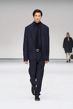 Placeholder for: A model in a dark suit and black shirt walks on a white runway. Another person is seen blurred in the background.
