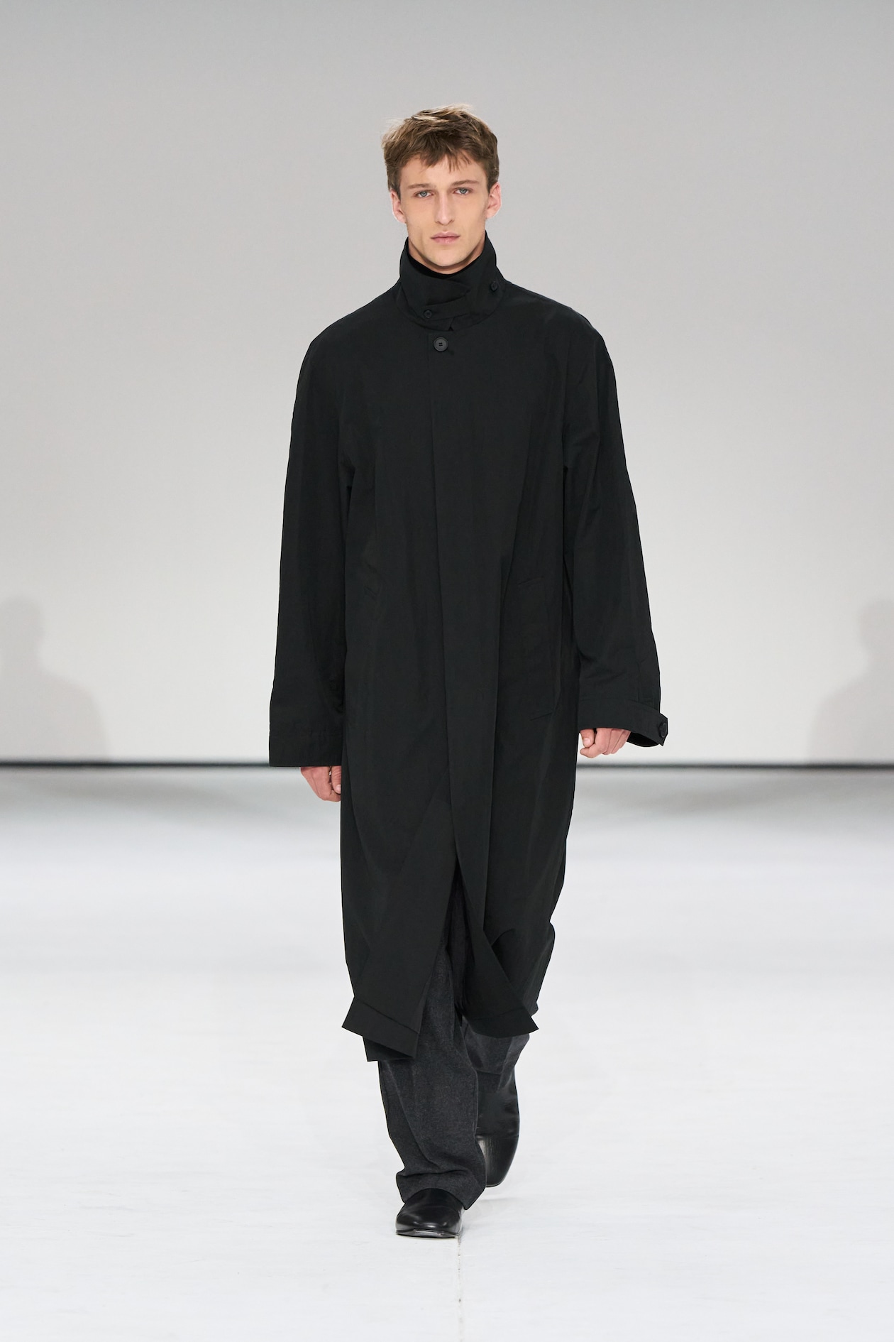 A person in a long black coat and dark pants walks confidently on a minimalist runway with a neutral background.