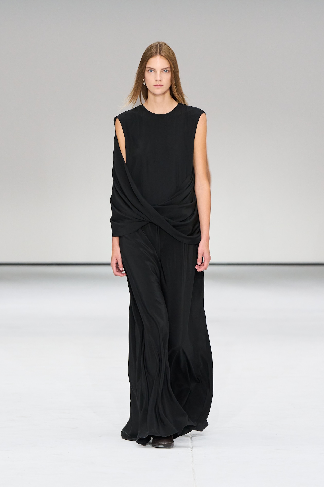 Model in a flowing black dress walks on a runway against a plain background, exuding elegance and confidence.