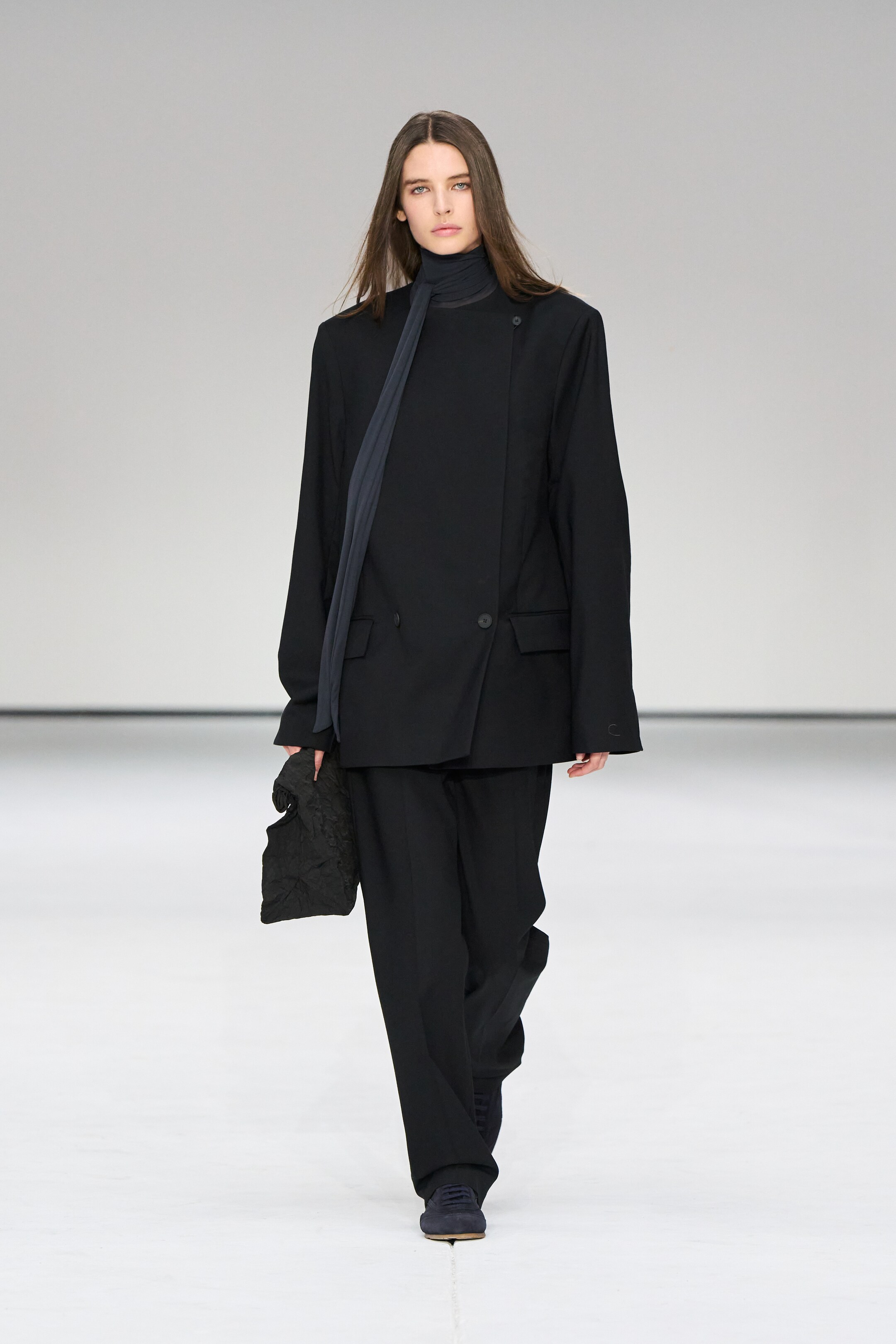 Model in an oversized black suit and scarf walks on a runway with a white background.