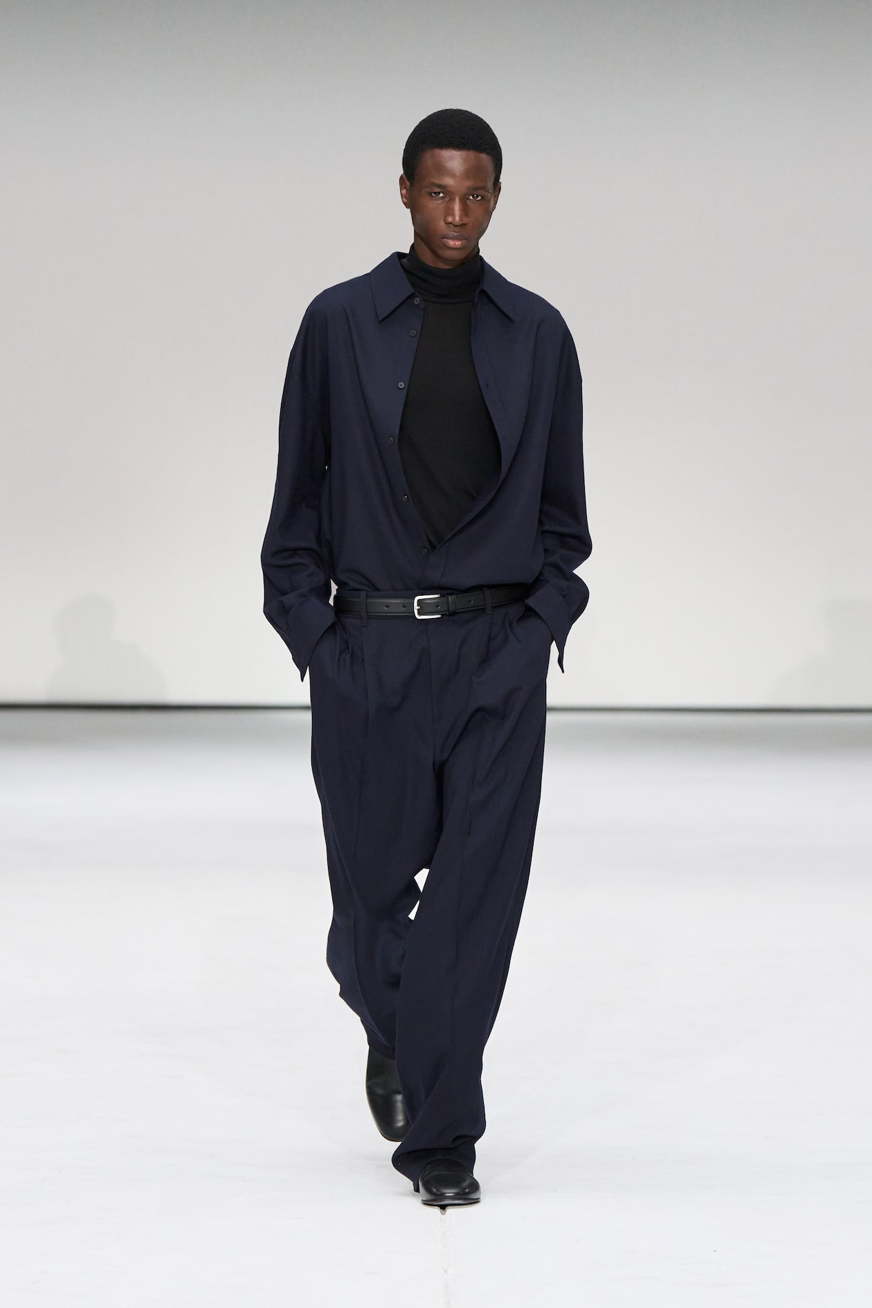 Model in a navy suit with a black turtleneck, walking on a white runway. Hands in pockets, confident expression.