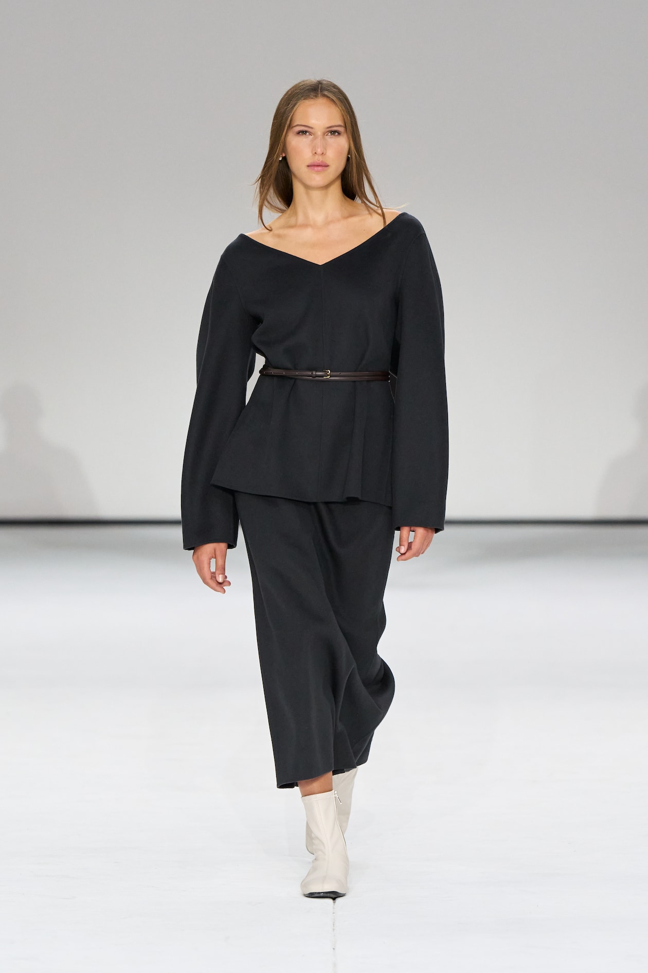 Model in a black V-neck outfit with a belt, walking on a runway, wearing white ankle boots against a plain background.