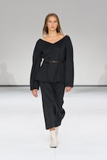 Placeholder for: Model in a black V-neck outfit with a belt, walking on a runway, wearing white ankle boots against a plain background.