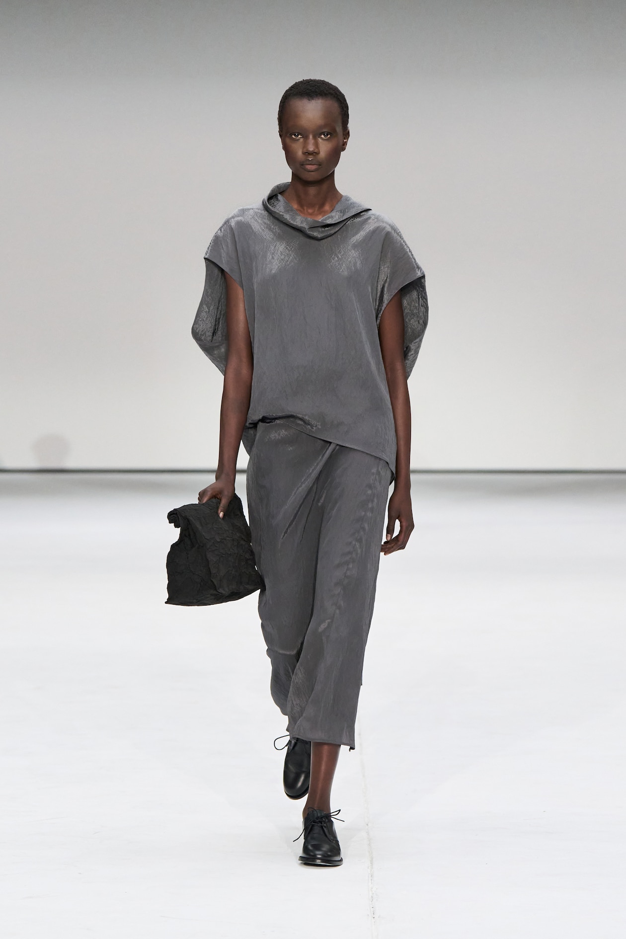 Model in a gray asymmetrical outfit and black shoes walks on a minimalist white runway, holding a textured black bag.