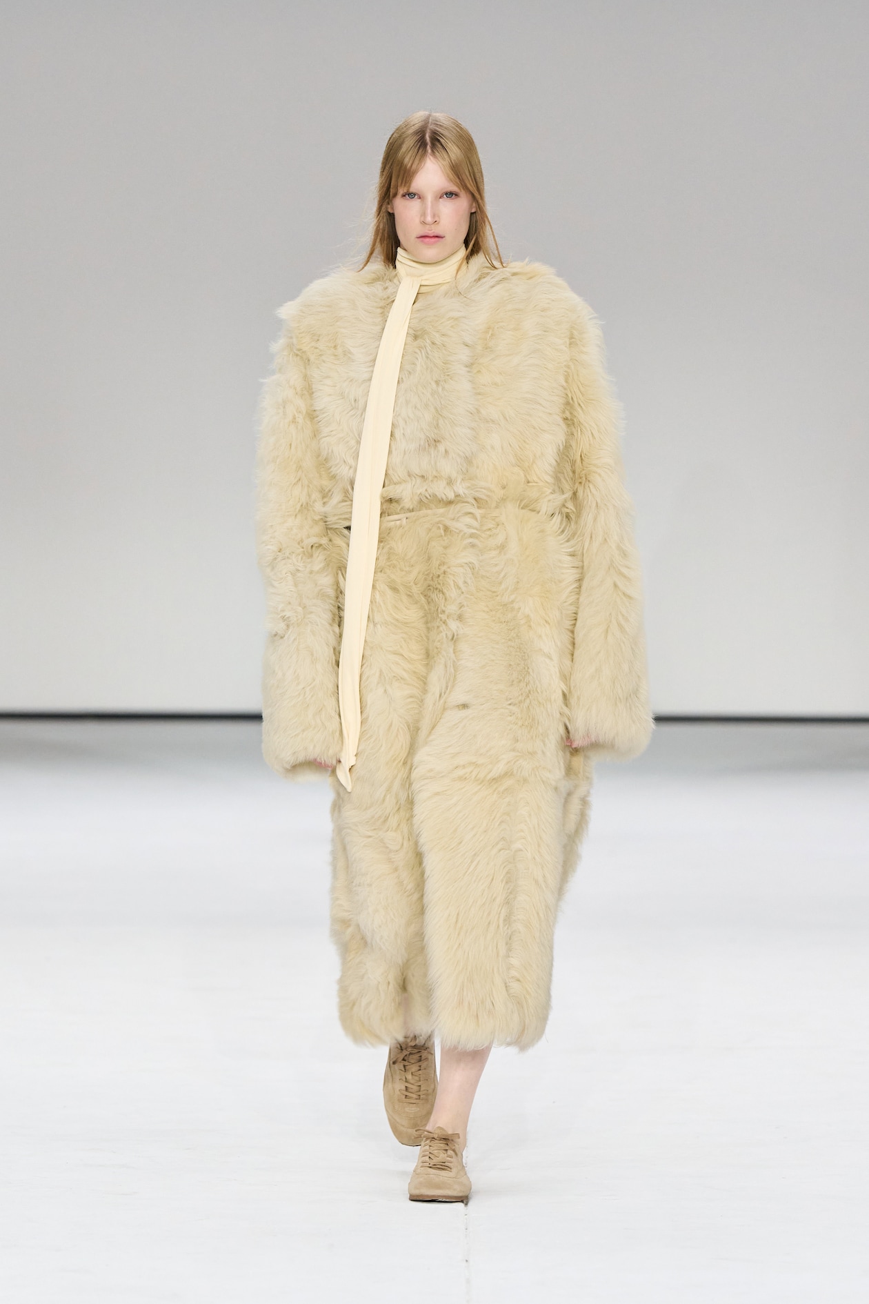 Model in a long, fluffy beige fur coat and matching scarf walks on a white runway, wearing tan shoes.