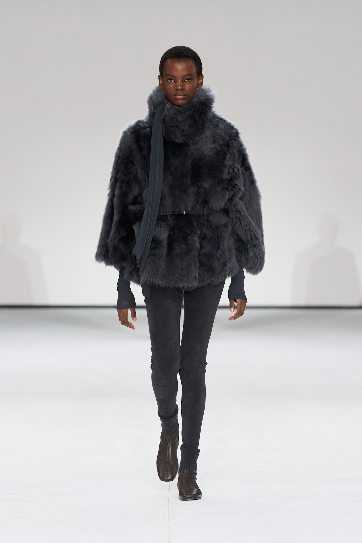 Model in dark, fluffy fur coat and scarf walks runway, wearing slim black pants and brown ankle boots against a plain white background.